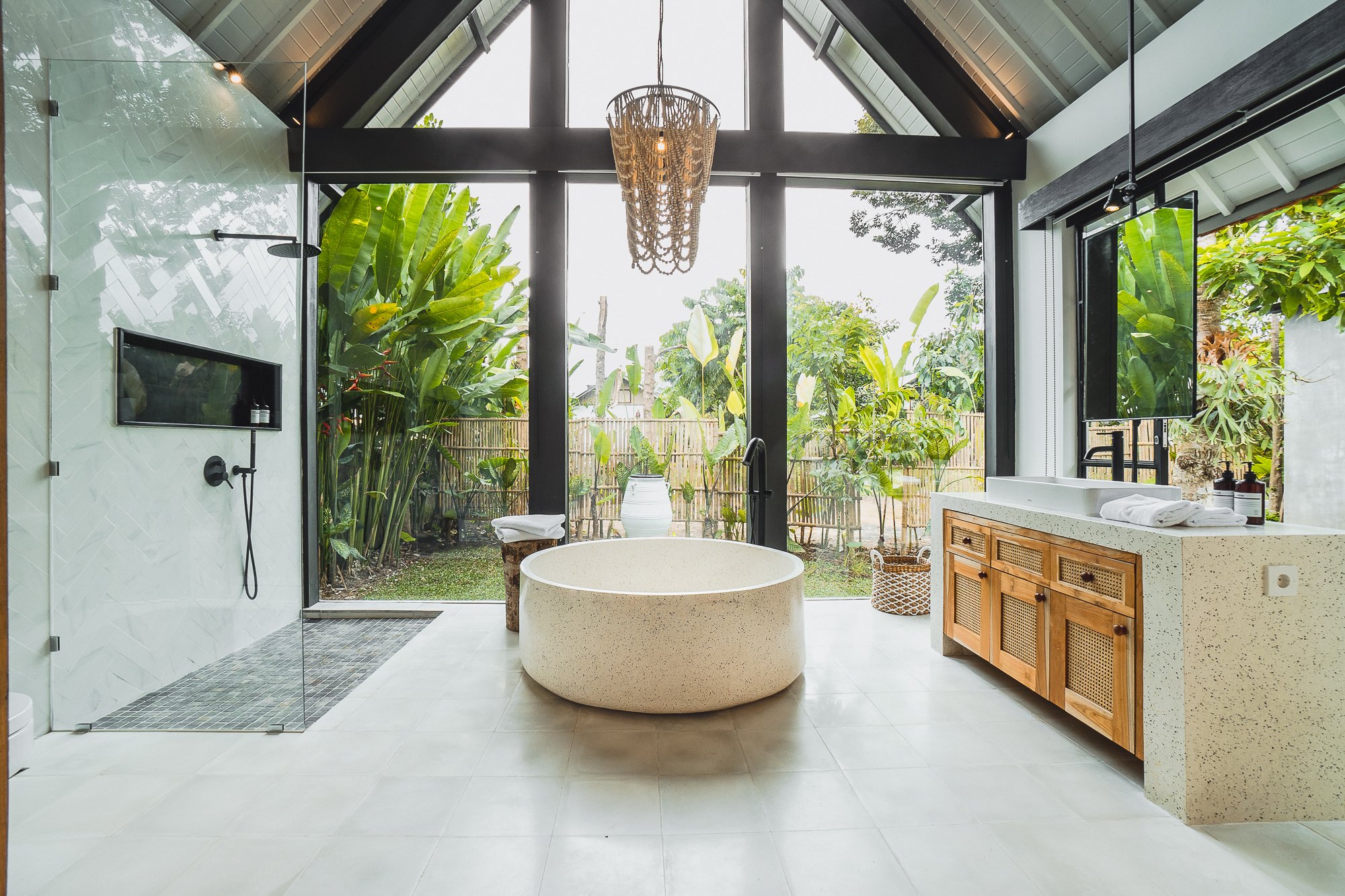 Modern bathroom with a freestanding bathtub, a rainforest shower, wooden vanity, and large glass windows overlooking lush greenery.