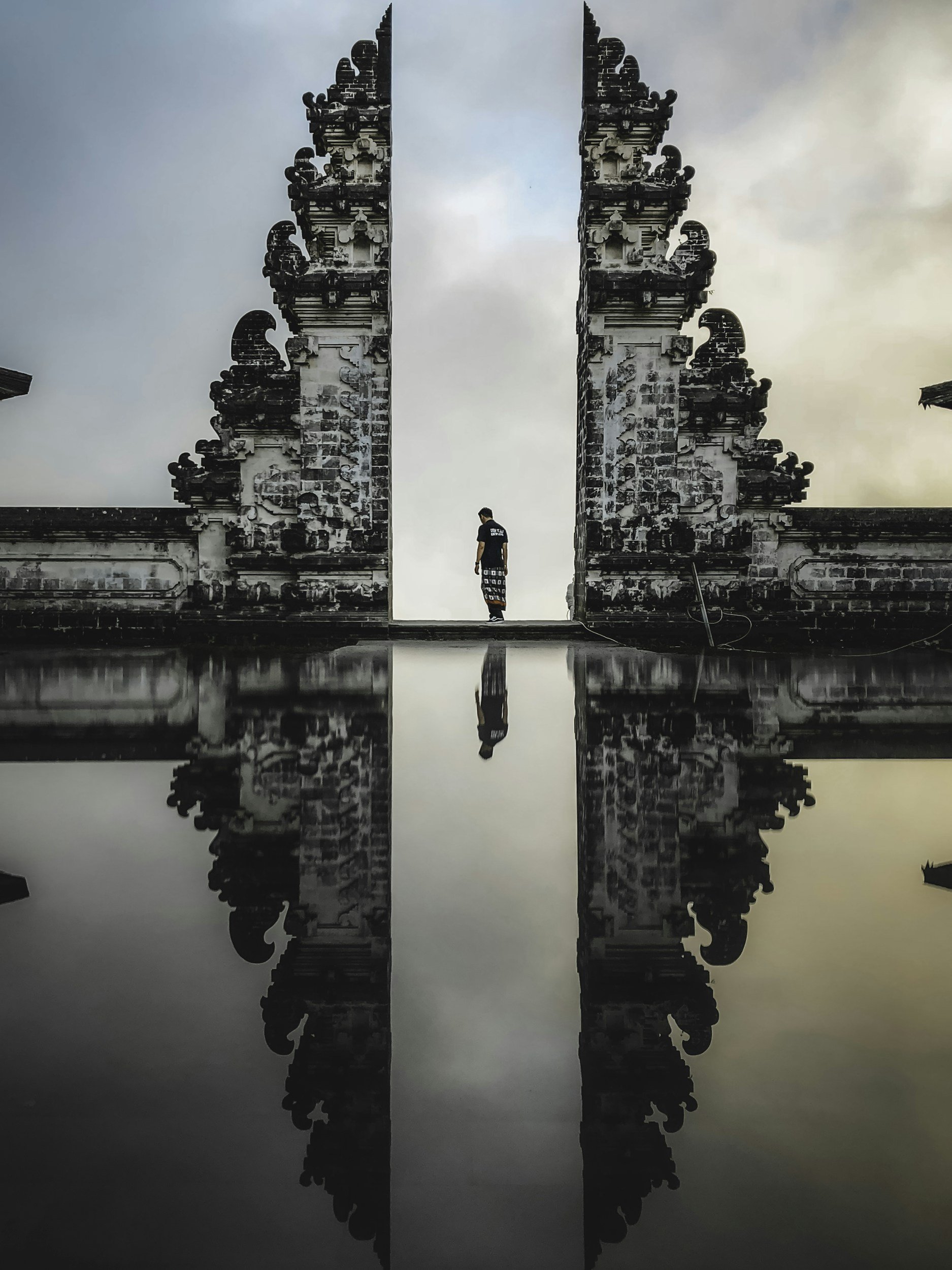 A person stands between two ornate stone pillars with intricate carvings, reflected in a still water surface, under a cloudy sky.