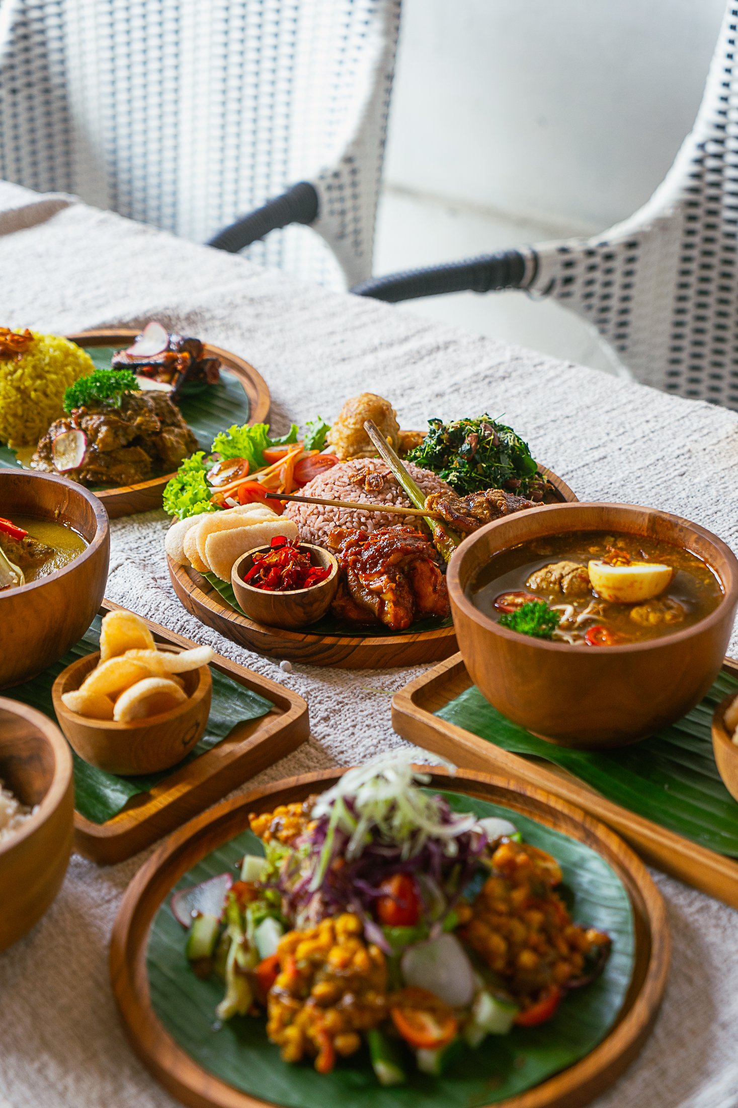 Assorted traditional Asian dishes served on wooden and ceramic plates, including rice, curry, soup, salad, and pickles, arranged on a table.