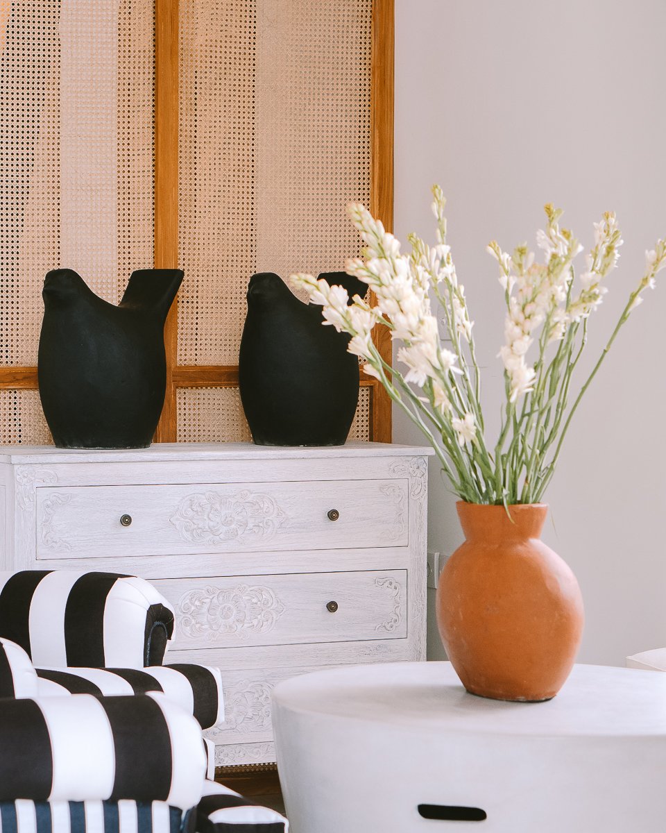 Room interior with a white dresser, two black vases on top, a striped black and white chair, and a large terracotta vase with white flowers on a white table.