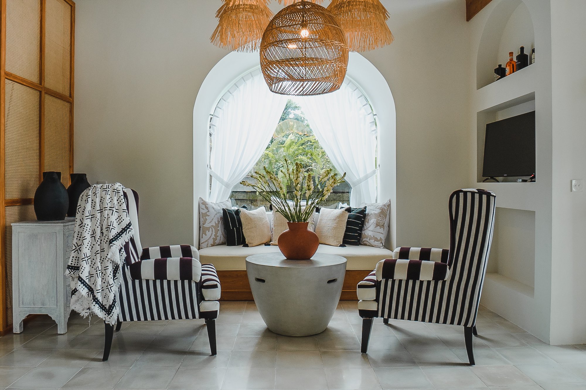 Living room with large arched window, white curtains, two striped armchairs, a round table with a vase of flowers, a built-in window seat with pillows, and a wall-mounted TV.