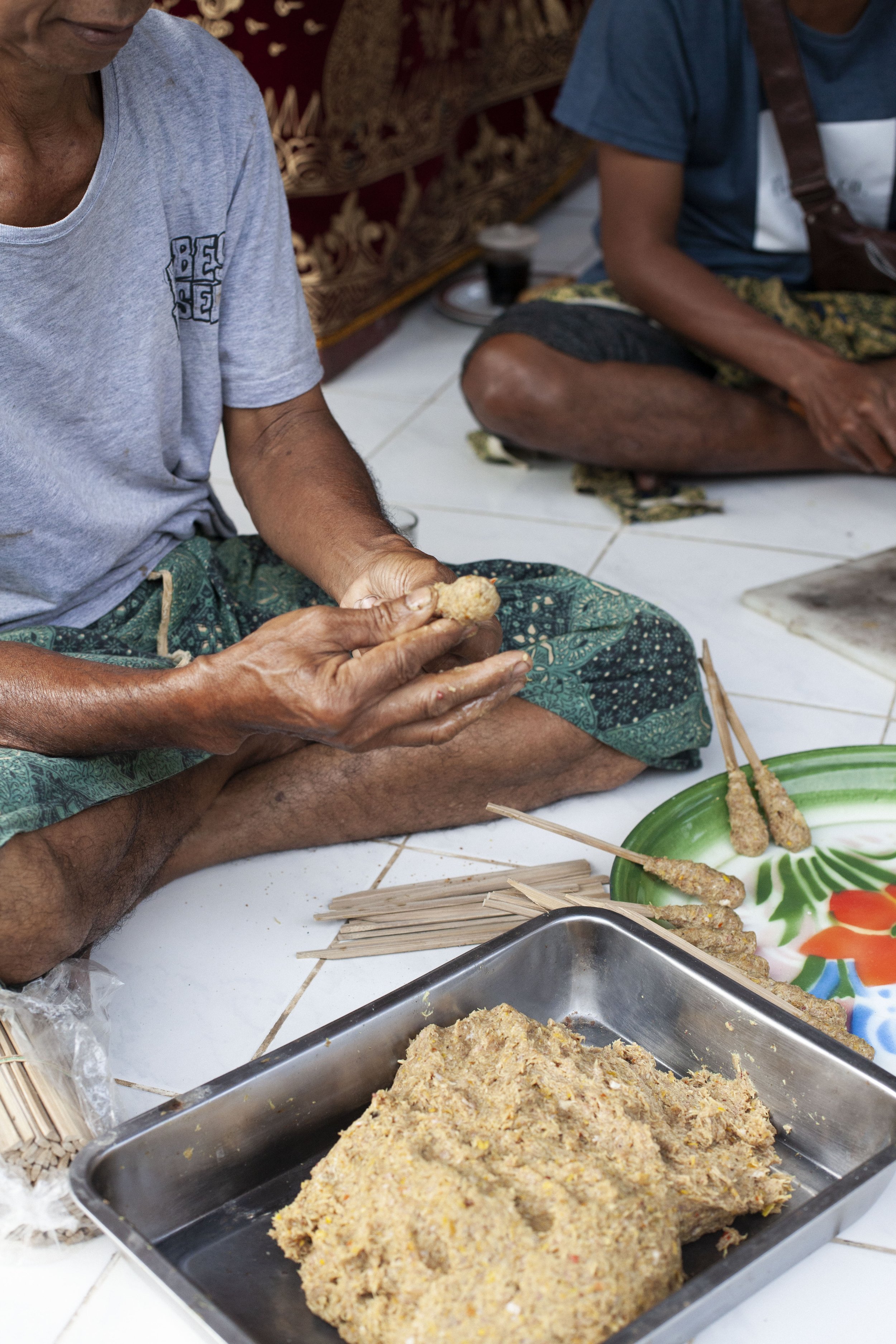 Two men sitting on the floor preparing satay skewers with a tray of ground peanut mixture and skewers nearby.