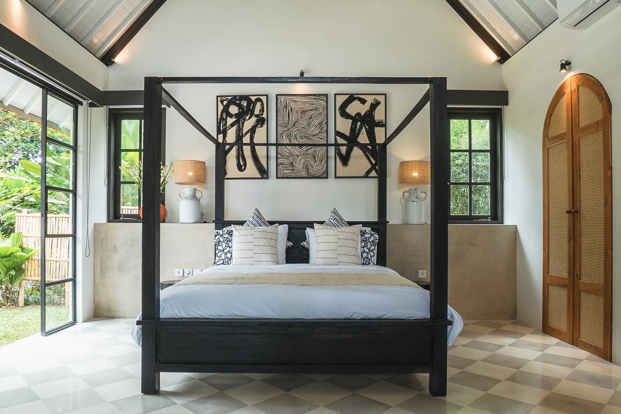 Bedroom with a black four-poster bed, white bedding, and patterned pillows, featuring abstract art on the wall, two bedside lamps, windows showing greenery, and wooden closet door.
