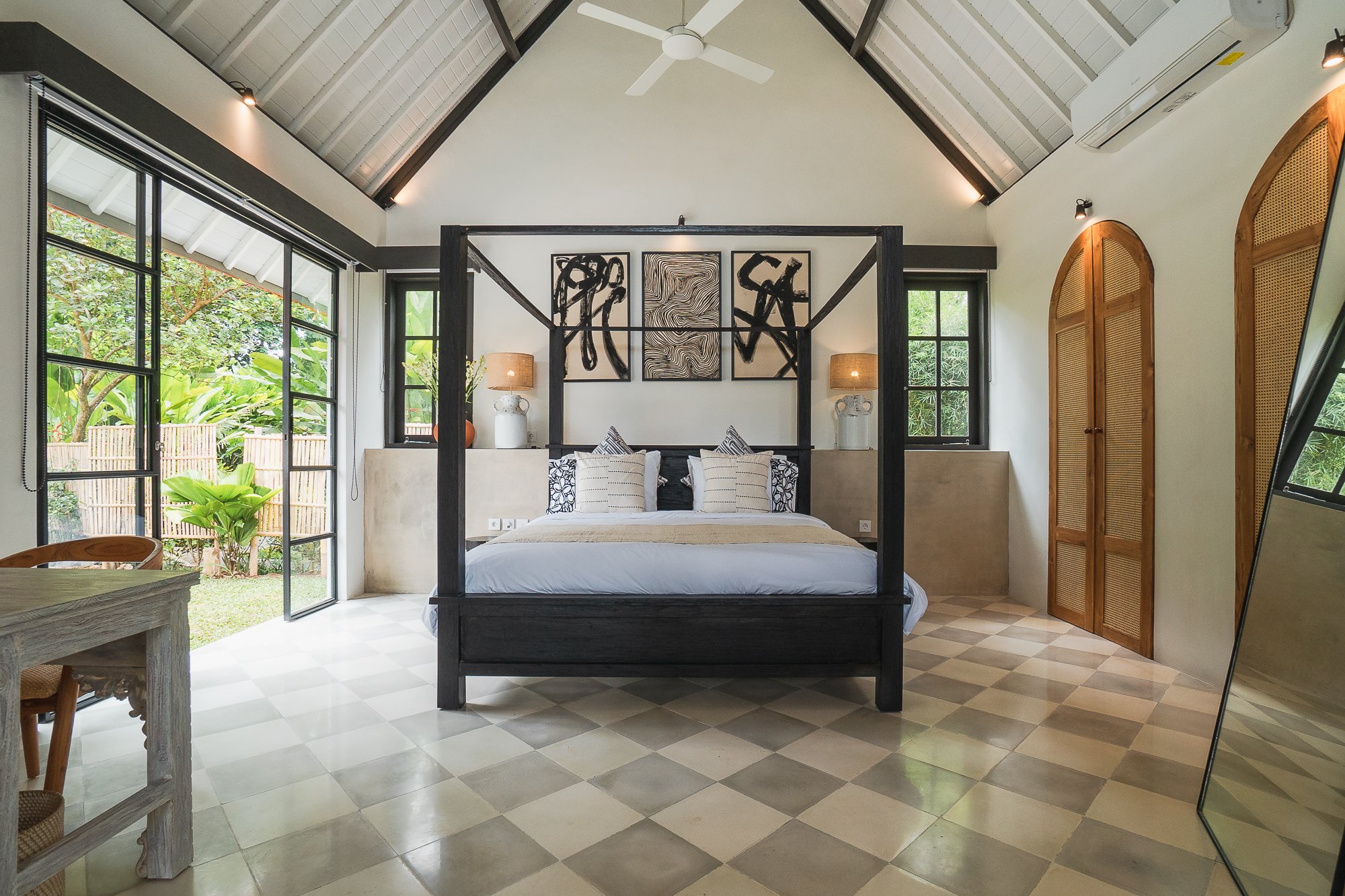 A tropical house with a tiled roof and large glass doors opens to a wooden patio with two lounge chairs by a small pool, surrounded by lush greenery and trees.